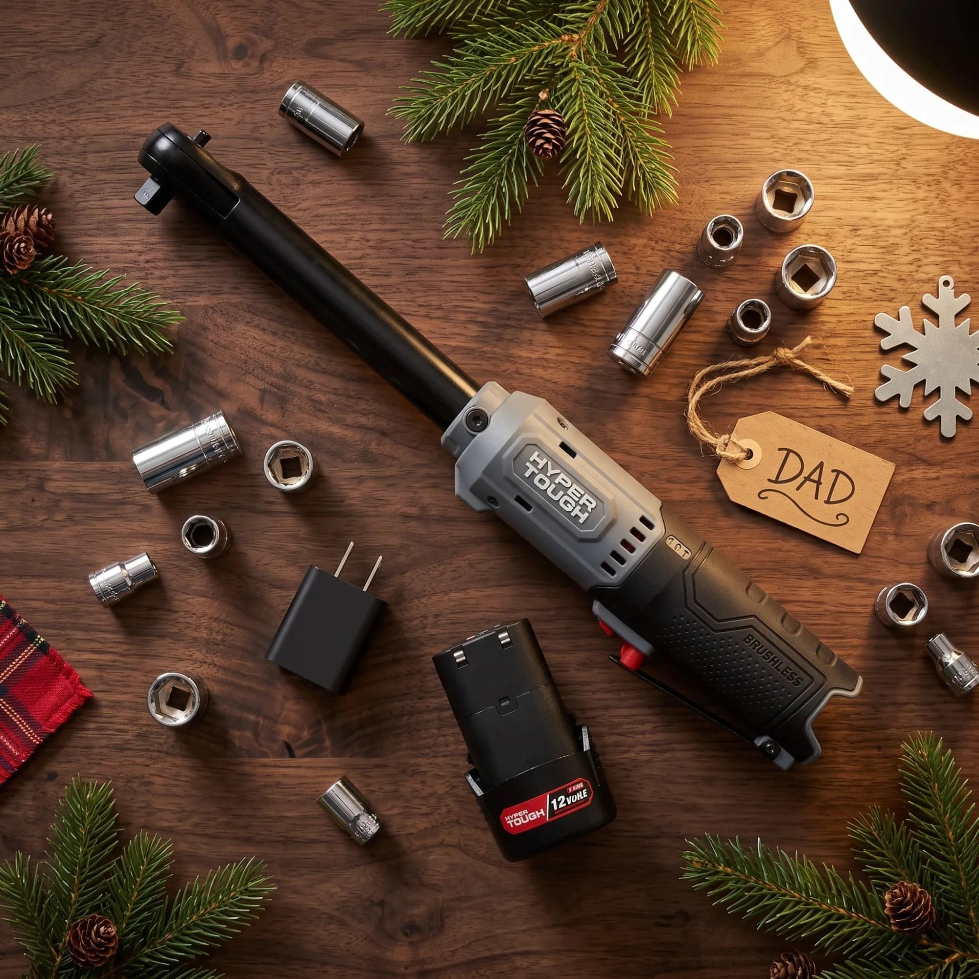 Holiday flat lay of the Hyper Tough extended reach ratchet wrench with its 12V battery, charger, and a ring of chrome sockets among pine branches, pinecones, and a gift tag reading Dad