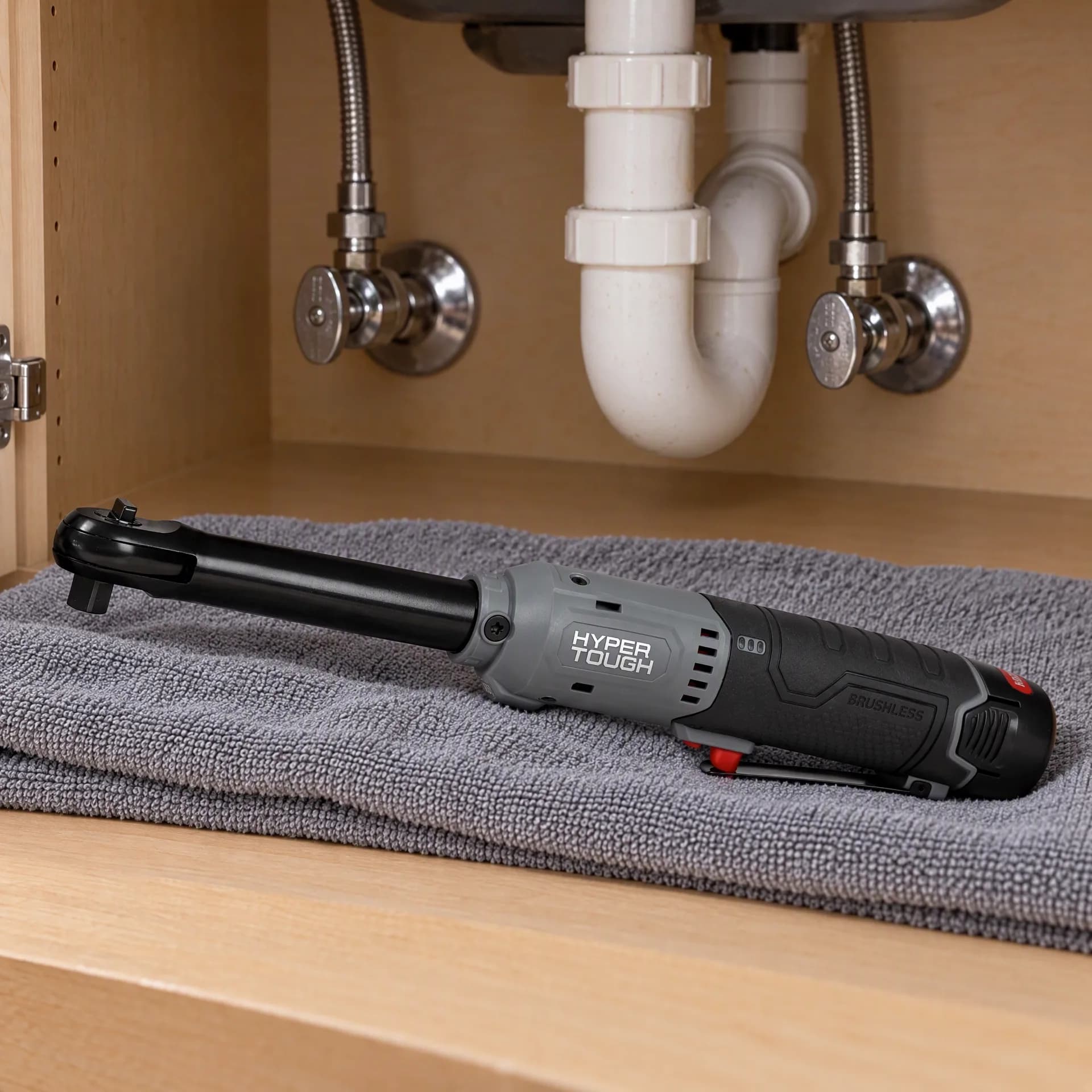 Hyper Tough extended reach ratchet wrench on a folded gray towel inside a cabinet under a kitchen sink, with the P-trap and supply valves above it